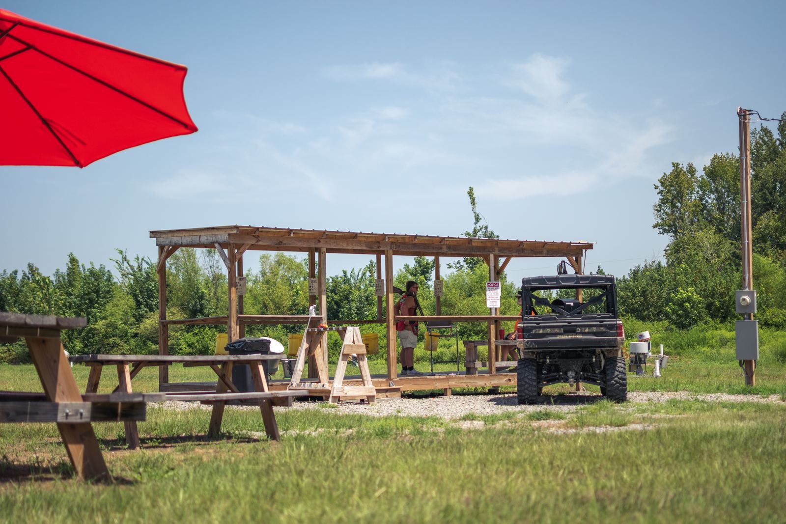 The covered shooting stand seen across the grass, with picnic tables and a red umbrella in the foreground and a utility vehicle parked alongside