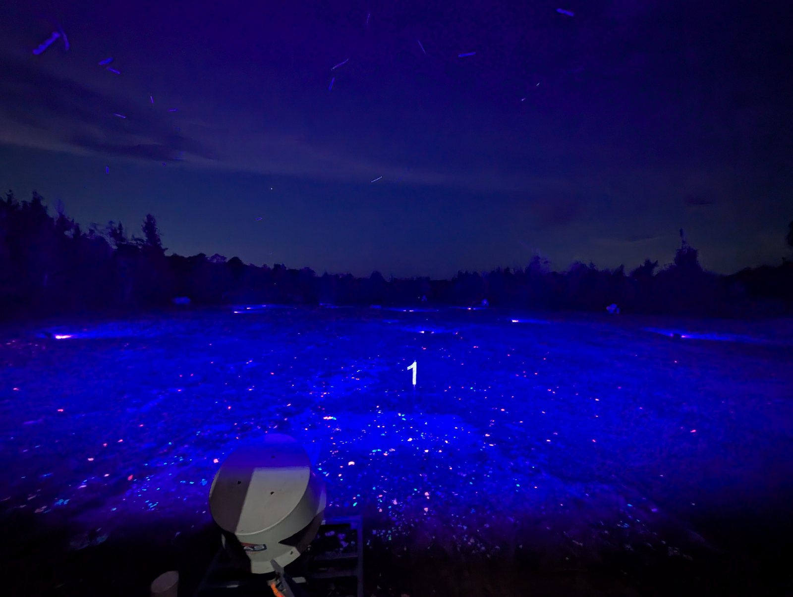 The field under blacklight, the ground and scattered clay fragments glowing blue, with a thrower in the foreground and broken pieces streaking across the twilight sky