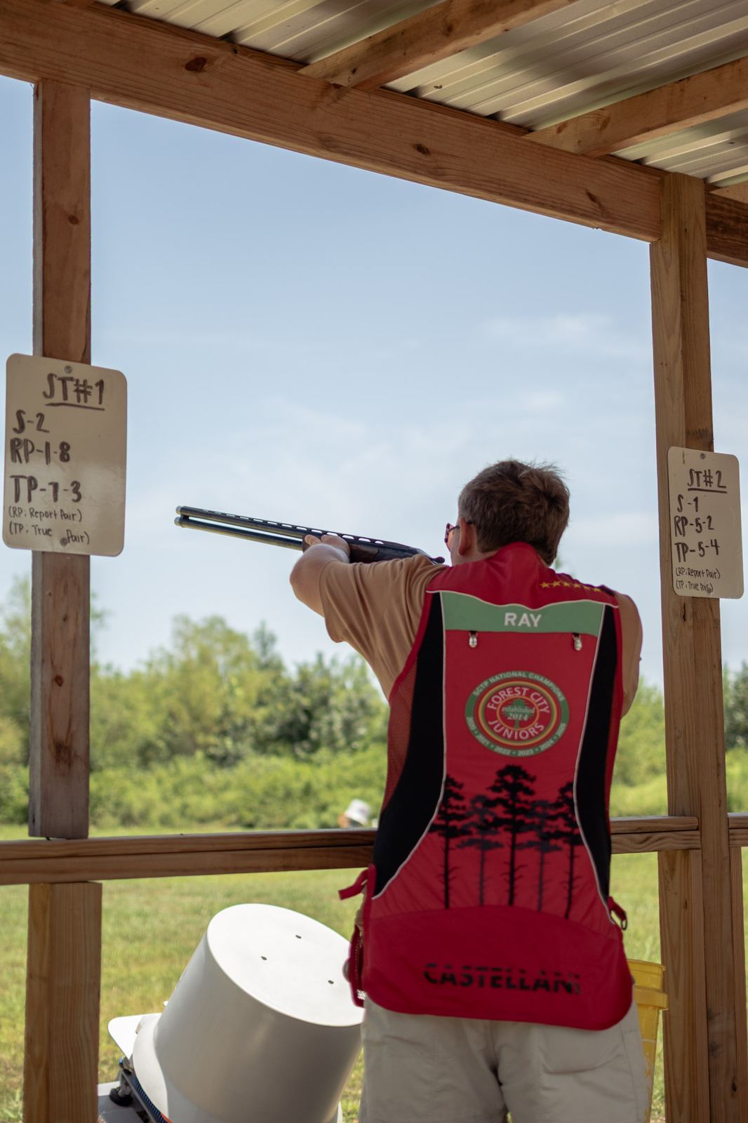 A shooter in a red vest mounted on a shotgun at station one, with the station menu sign posted on the timber beside him